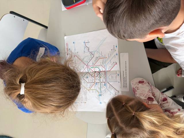 Drei Kinder beugen sich über einen Tisch und betrachten eine U-Bahn-Karte und Aufgabenblätter.
