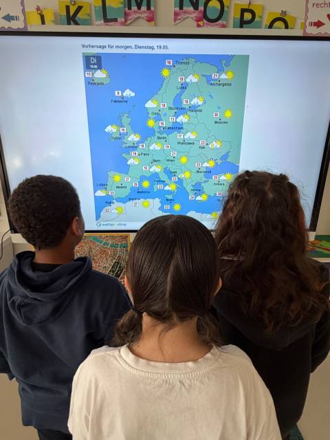 Drei Kinder betrachten von hinten eine Wetterkarte von Europa auf einem großen Bildschirm.