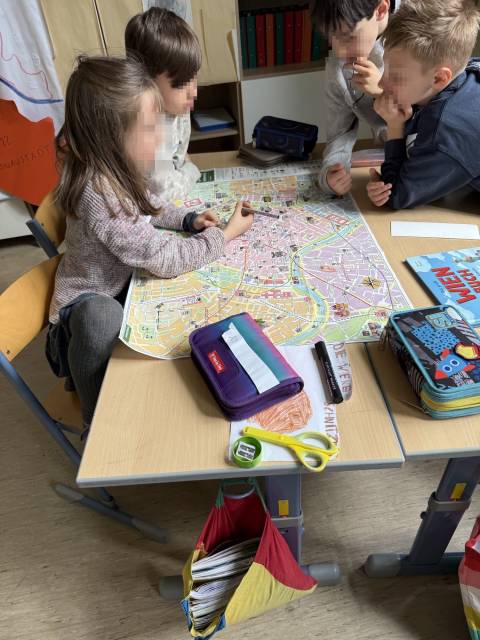 Vier Kinder beugen sich über eine große Stadtkarte auf einem Tisch, ein Mädchen zeigt mit einem Stift darauf.