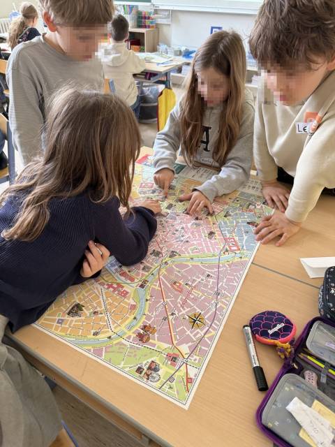 Vier Kinder beugen sich über eine große Stadtkarte auf einem Schreibtisch und zeigen auf verschiedene Orte.