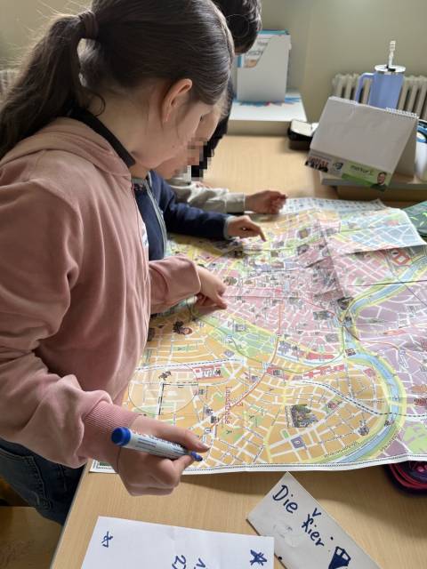 Zwei Kinder studieren eine große Stadtkarte auf einem Tisch, zeigen darauf und halten einen blauen Marker.