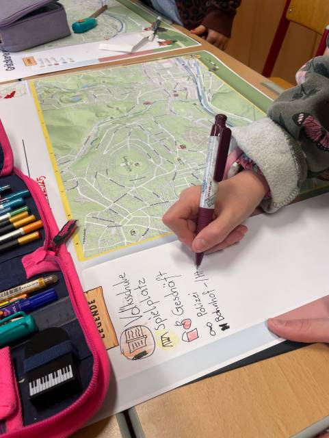 Eine Kinderhand schreibt mit einem Stift auf einem Blatt mit einer Kartenlegende neben einer Stadtkarte.