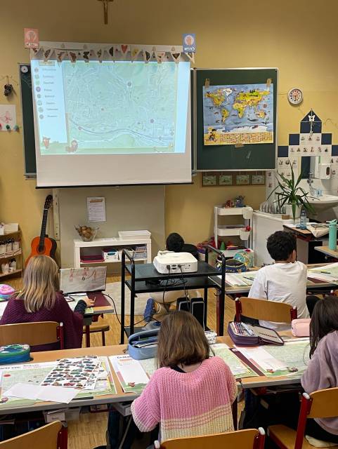 Schüler sitzen in einem Klassenzimmer, blicken auf eine projizierte Stadtkarte und arbeiten an ihren Tischen.